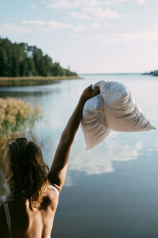 Person holding a pillow by a lake with trees in the background